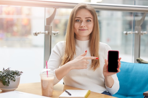 Słodka Blond Włosy Kobieta Siedzi W Kawiarni, Trzymając Smartfon