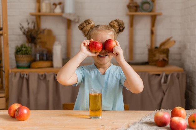 Słodka dziewczyna trzyma szklankę z sokiem i jabłkiem, siedząc przy drewnianym stole
