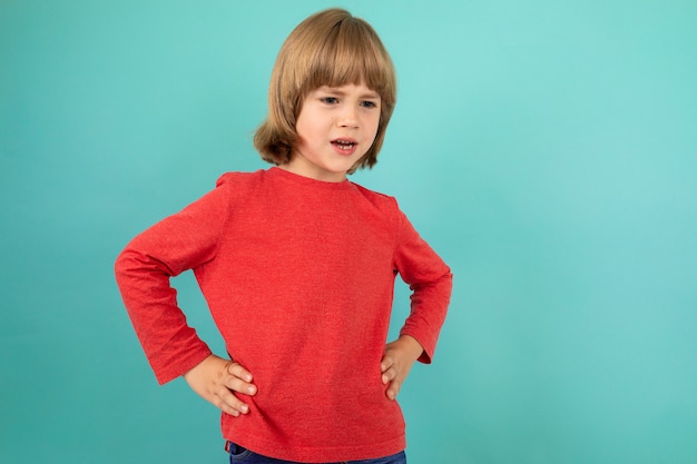 Słodka europejska chłopiec w czerwonym pulowerze pozuje na odosobnionym błękitnym studiu