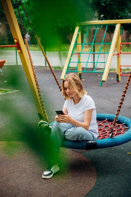 Słodka nastolatka trzymająca telefon w dłoni siedzi na huśtawce w nowoczesnym parku Uczennica pisze wiadomości do znajomych online Komunikując się w sieciach społecznościowych, blogując przez telefon