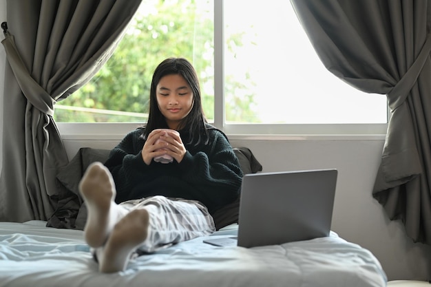 Słodka nastolatka w ciepłym swetrze pijąca gorącą czekoladę i używająca laptopa w sypialni