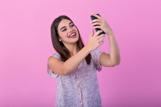 Słodka piękna dziewczyna z długimi włosami w studiu z różowymi ścianami robi selfie Patrzy na swój smartfon i robi selfie