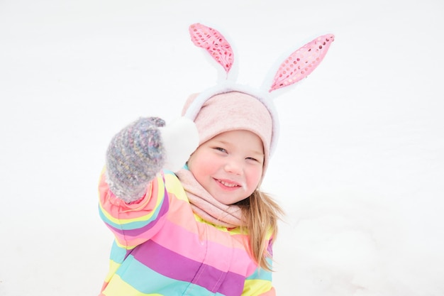 Słodka, wesoła przedszkolanka z uszami królika na głowie bawi się sercem wykonanym ze śniegu w kwietniu