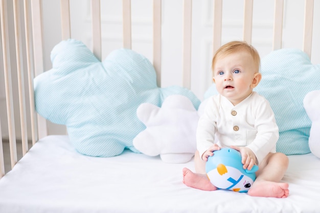 Słodki blond chłopiec w wieku 6 miesięcy w łóżeczku w jasnej sypialni w białym bawełnianym body z niebieską zabawką koncepcja towarów dla dzieci