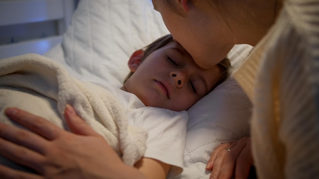Zdjęcie słodki chłopczyk śpi, podczas gdy troskliwa matka całuje go i poprawia koc