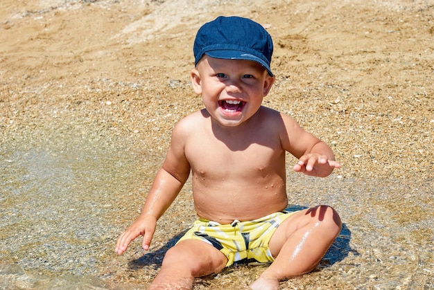 Słodki chłopczyk w niebieskiej czapce uśmiecha się siedząc na mokrym piasku plaży w nadmorskim miasteczku Omis