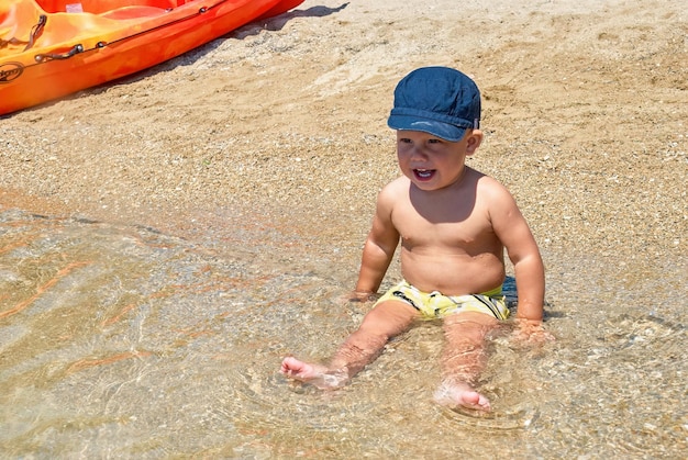 Słodki chłopczyk w niebieskiej czapce uśmiecha się siedząc na mokrym piasku plaży w nadmorskim miasteczku Omis