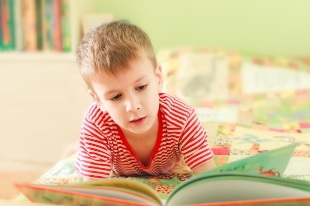 Zdjęcie słodki chłopiec czyta książkę z alfabetem, leżąc na łóżku. dziecko patrzy na otwartą książkę.