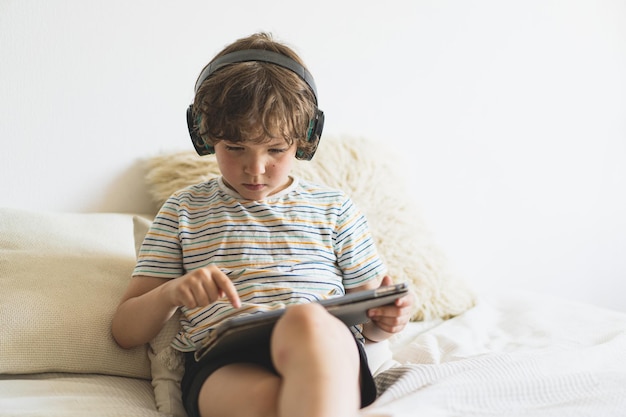 Słodki chłopiec leżący na łóżku w domu i bawiący się tabletem PC lub czytający online i słuchający muzyki przez bezprzewodowe słuchawki