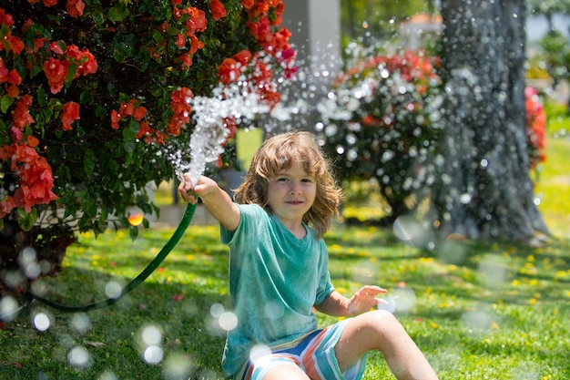 Słodki chłopiec podlewa rośliny w ogrodzie w letni dzień Dziecko z narzędziami ogrodniczymi i wężem do podlewania w przydomowym ogródku Dziecko bawi się na podwórku Pojęcie dobroci dziecka i dzieciństwa