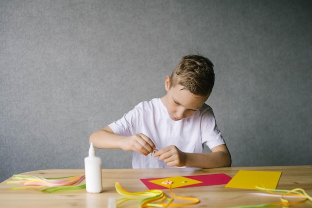 Słodki chłopiec robi kwiaty na pocztówkę, wykonując quilling, siedząc przy stole