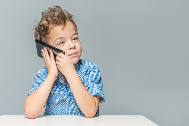 Słodki chłopiec rozmawiający przez telefon, siedząc przy stole na szarym tle.