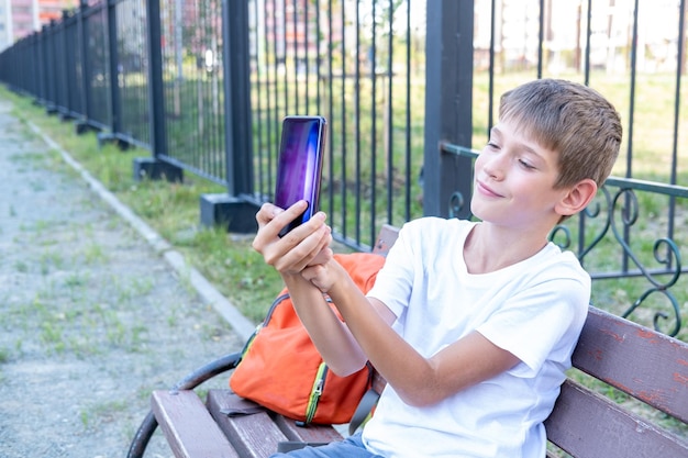 Słodki chłopiec uczeń używa telefonu robi selfie komunikuje się przez wideorozmowę z przyjaciółmi siedzącymi na ławce