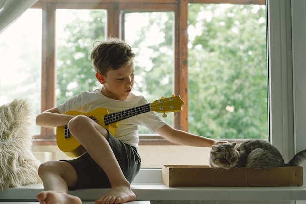Słodki chłopiec uczy się grać na gitarze Ukulele na parapecie w pobliżu okna z kotem Przytulny dom Letnie wakacje styl życia