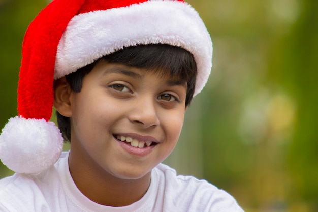 Słodki chłopiec w czapce Mikołaja i białej koszulce, uśmiechający się radośnie i patrzący w kamerę