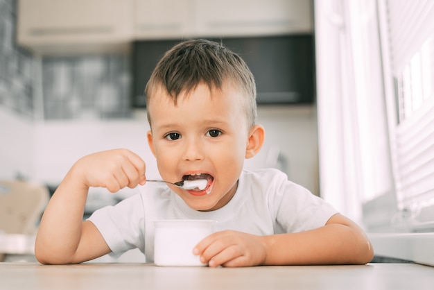 Słodki chłopiec w kuchni je jogurt w lekkich ubraniach z pojemnika na biały jogurt, miejsce na reklamę