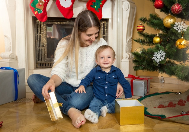 Słodki chłopiec z matką otwierają pudełka z prezentami pod choinką w salonie