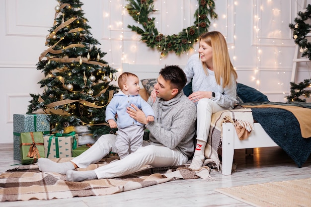 Zdjęcie słodki chłopiec ze szczęśliwymi rodzicami przy choince rodzina z małym dzieckiem w sypialni przy udekorowanej choince świąteczna atmosfera nowego roku rodzinne świętowanie