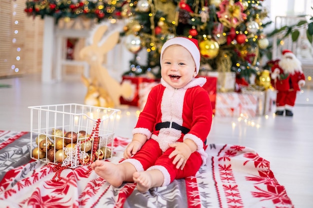 Słodki mały chłopiec w stroju Świętego Mikołaja siedzi z zabawkami choinkowymi pod świąteczną choinką w domu szczęśliwe dziecko świętuje Boże Narodzenie i Nowy Rok w domu