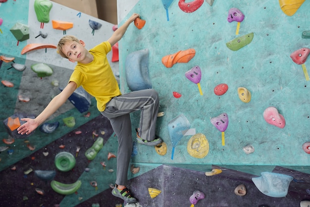 Słodki młodzik w żółtej koszulce i szarych spodniach patrzy na Ciebie trzymając małą skałę na ściance wspinaczkowej podczas ćwiczeń
