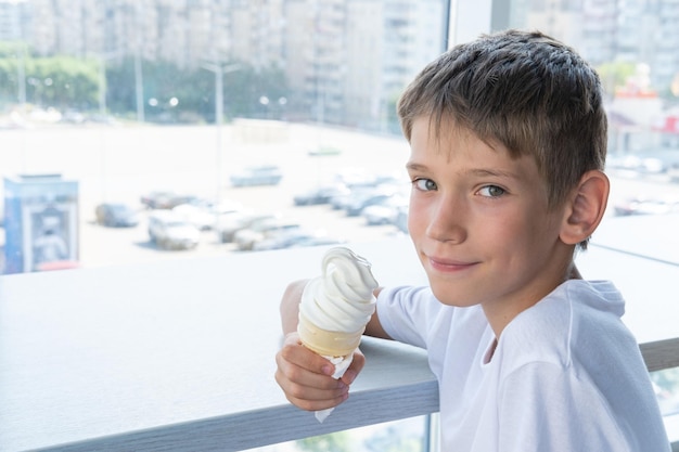 Słodki nastoletni chłopak je wirujące białe lody w kubku waflowym siedząc przy stoliku przy oknie w kawiarni kopia przestrzeni Niewyraźne tło Dziecko lubi lody Słodka przekąska