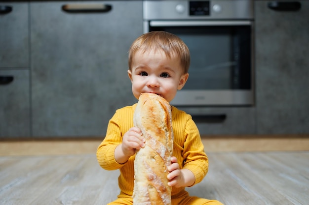 Słodki roczny chłopiec siedzi w kuchni i je długi chleb lub bagietkę w kuchni Pierwsze zjedzenie chleba przez dziecko Chleb jest dobry dla dzieci