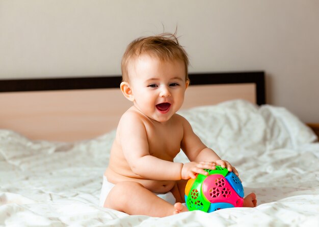 Słodkie blond dziecko bawi się piłką, siedząc na łóżku w sypialni. szczęśliwe dziecko w wieku 6 miesięcy bawiące się piłką