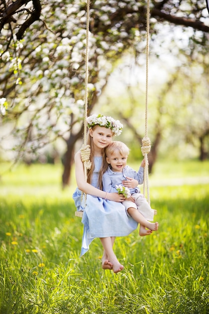 Słodkie dziewczyny i chłopca, zabawy na huśtawce w kwitnącym starym ogrodzie jabłoni. Słoneczny dzień. Wiosenne zajęcia na świeżym powietrzu dla dzieci