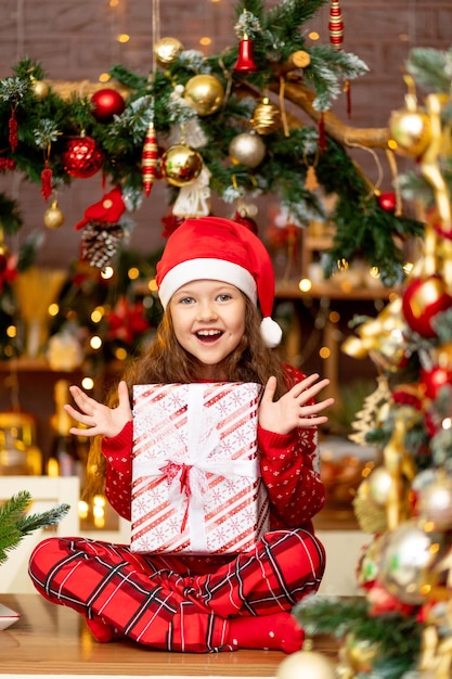 Słodkie małe dziecko dziewczynka w zimowe ubrania Czapka Świętego Mikołaja i czerwony sweter raduje się prezentem i śmieje się ze szczęścia w ciemnej kuchni domu koncepcja nowego roku i Bożego Narodzenia