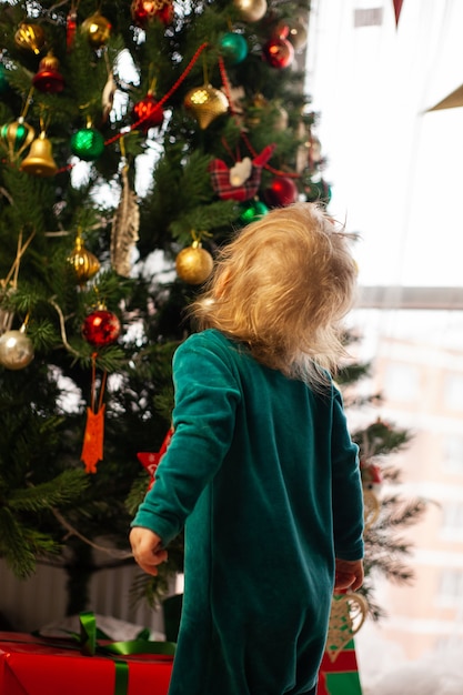 Słodkie małe dziecko patrzy na choinkę ozdobioną zabawkami i girlandą. Nowy rok i Boże Narodzenie. Ferie. Komfort w domu. Zimowe wakacje styl życia. Szczęśliwe dzieciństwo.