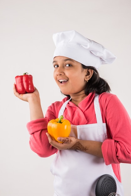 Słodkie małe indyjskie dziecko lub dziewczynka przebrana za szefa kuchni i trzymająca naczynia do gotowania lub warzywa, stojąca na białym tle nad białym tłem