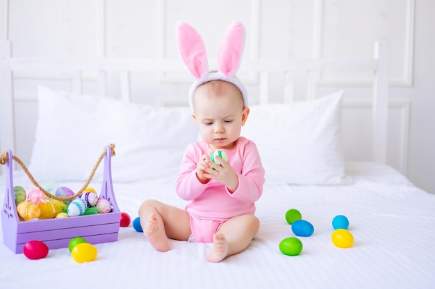 Słodkie śmieszne dziecko z uszami królika z kolorowymi pisanki i koszem jaj w domu na łóżku z białej pościeli Wielkanocna kartka z życzeniami