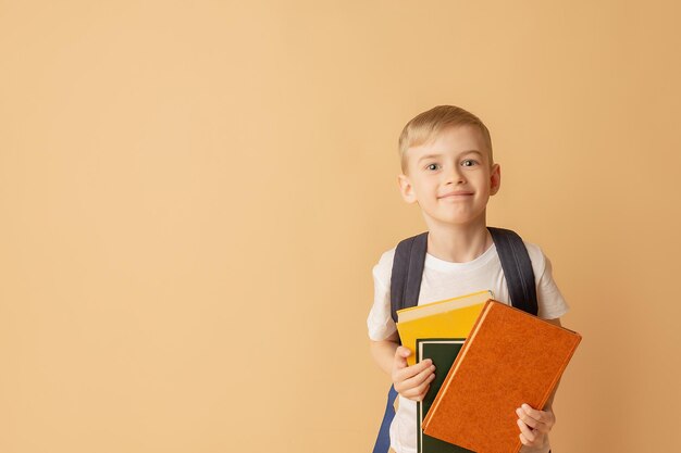 Słodkie uśmiechnięte dziecko z plecakiem trzymające książki w ramach przygotowań do szkoły na beżowym tle