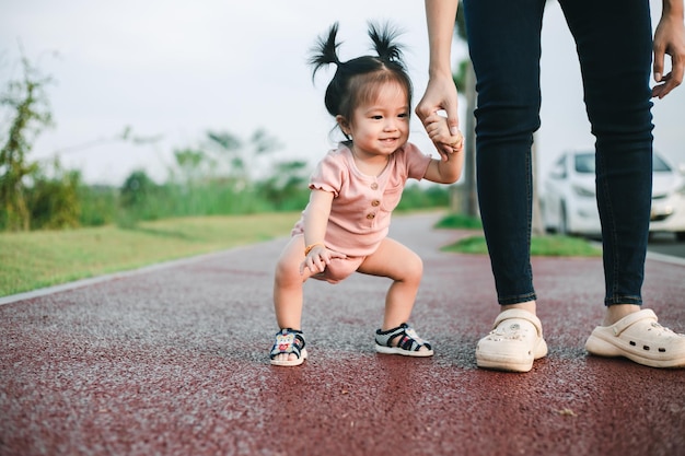 Słodkie, zabawne, szczęśliwe dziecko robi swoje pierwsze kroki w parku, matka trzyma ją za ręce, wspierając ją, ucząc się chodzić.