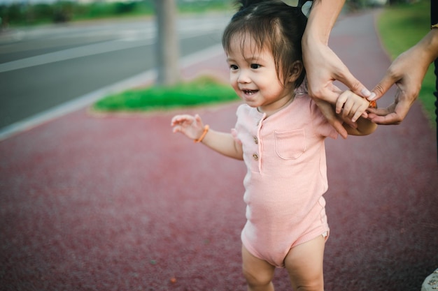 Słodkie, zabawne, szczęśliwe dziecko robi swoje pierwsze kroki w parku, matka trzyma ją za ręce, wspierając ją, ucząc się chodzić.
