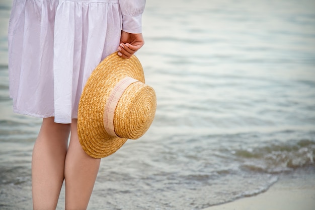 Słomkowy kapelusz w kobiecej dłoni nad brzegiem morza o zachodzie słońca. Nie do poznania kobieta w białej sukni plaży po odpoczynku o zachodzie słońca na plaży. Kobieta cieszy się wakacjami i wolnością na plaży o zachodzie słońca. Skopiuj miejsce.