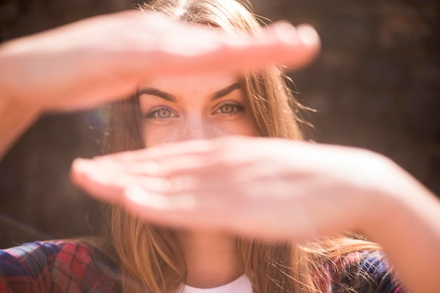 Słoneczny portret pięknej modelki rasy kaukaskiej z niebieskimi oczami w podświetleniu z rękami przed jej twarzą, aby ukryć niewyraźne tło bokeh w ustach