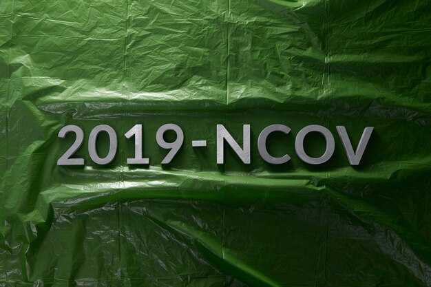 Słowo 2019NCOV ułożone metalowymi literami na zielonym tle zmiętej folii polietylenowej w płaskiej kompozycji