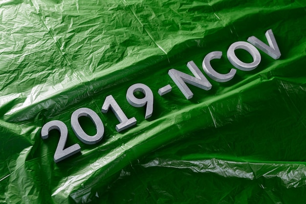 Słowo 2019NCOV ułożone metalowymi literami na zielonym tle zmiętej folii z tworzywa sztucznego w ukośnej kompozycji ukośnej