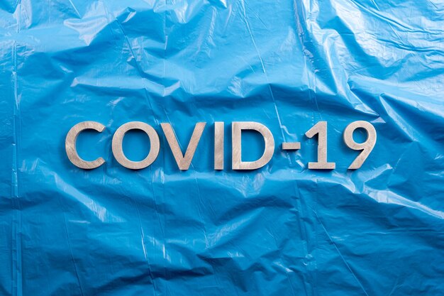 Słowo COVID19 ułożone z aluminiowymi literami na zmiętym niebieskim tle folii z tworzywa sztucznego w płaskiej kompozycji leżącej bezpośrednio nad widokiem