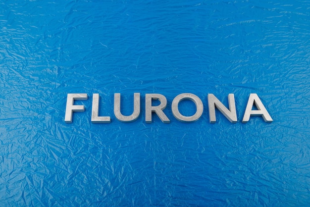 Słowo flurona ułożone srebrnymi metalowymi literami na zmiętym niebieskim tle folii z tworzywa sztucznego