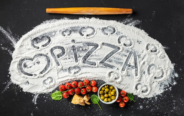 Słowo pizza i serce jest napisane na mące z wałkiem do ciasta i składnikami do robienia włoskiej pizzy, widok z góry. Abstrakcyjne tło do pieczenia