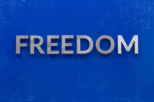 Słowo wolność ułożone srebrnymi metalowymi znakami na pomalowanej na niebiesko drewnianej desce w centralnej, płaskiej kompozycji