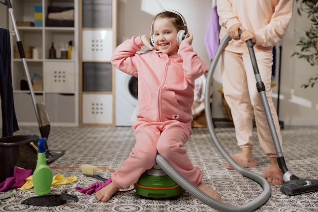 Słuchanie muzyki na słuchawkach bezprzewodowych podczas sprzątania łazienki podczas prac domowych mała dziewczynka siedzi na odkurzaczu, którego mama używa do czyszczenia podłogi
