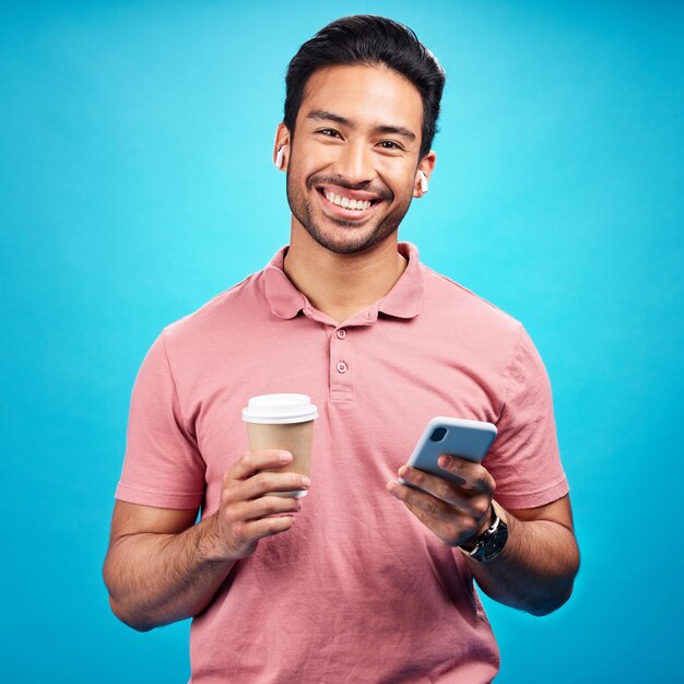 Słuchawki do kawy i portret mężczyzny z telefonem w studio na białym tle na niebieskim tle Telefon komórkowy z herbatą i szczęście azjatyckiej osoby z napojem kofeinowym i telefonem komórkowym do muzyki lub radia w mediach społecznościowych