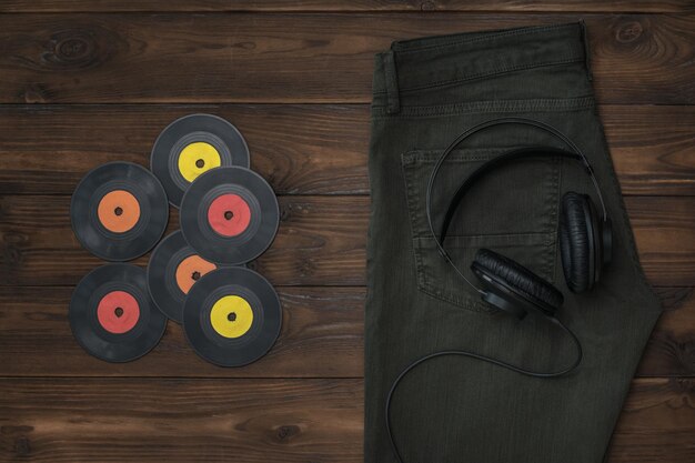 Słuchawki kolorowe płyty winylowe i zielone dżinsy na ciemnym drewnianym tle Pojęcie mody i muzyki
