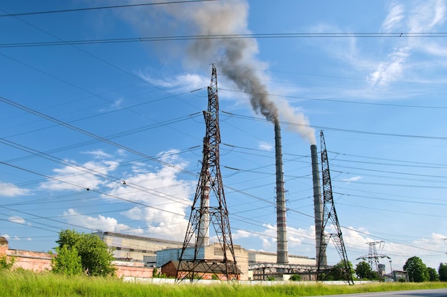 Słupy elektryczne wysokiego napięcia na wysokich rurach elektrowni węglowej z czarnym dymem unoszącym się w górę, zanieczyszczając atmosferę. Produkcja energii elektrycznej z wykorzystaniem koncepcji paliw kopalnych