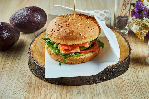 Smaczny burger z łososiem, pomidorami, rukolą i ogórkami na drewnianej desce na drewnianym stole. Burger rybny. Zdrowa przekąska. Zamknąć widok