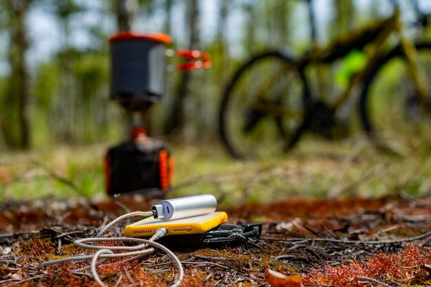 Smartfon jest ładowany przez power bank w lesie na tle turystycznego palnika gazowego i roweru