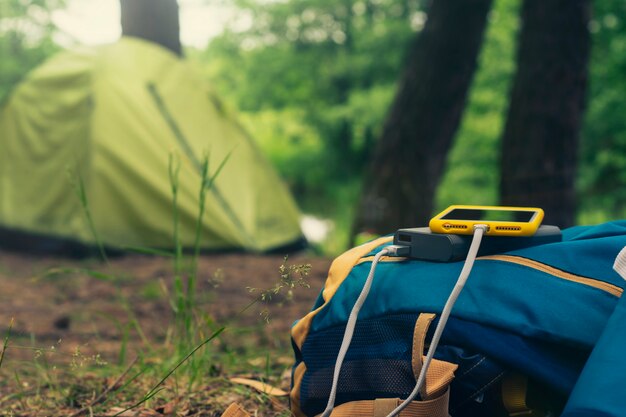 Smartfon jest ładowany za pomocą przenośnej ładowarki. Power Bank ładuje telefon na zewnątrz z plecakiem na tle namiotu.